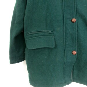 Puede incluir: Primer plano de un abrigo de lana verde oscuro. El abrigo tiene un bolsillo con cremallera, botones marrones y un dise&ntilde;o cl&aacute;sico. El abrigo est&aacute; hecho de un material grueso y c&aacute;lido, perfecto para el fr&iacute;o.