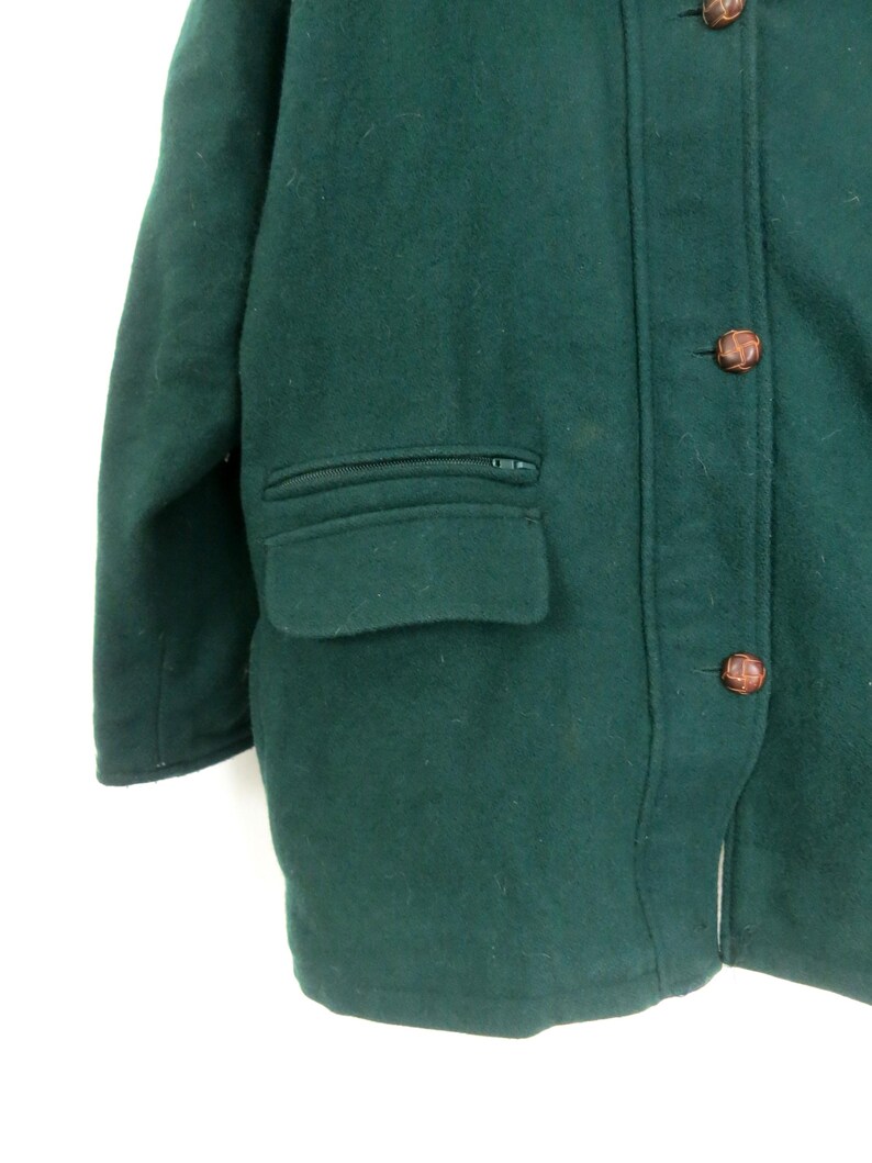 Puede incluir: Primer plano de un abrigo de lana verde oscuro. El abrigo tiene un bolsillo con cremallera, botones marrones y un dise&ntilde;o cl&aacute;sico. El abrigo est&aacute; hecho de un material grueso y c&aacute;lido, perfecto para el fr&iacute;o.