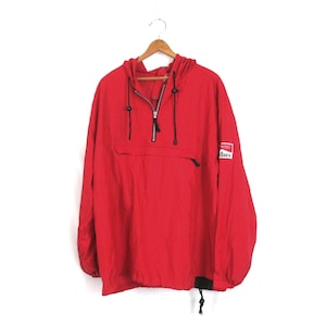 May include: A red anorak-style jacket with a hood, a half-zip closure, and a front pocket. The jacket has black drawstrings and a small patch on the sleeve. The jacket is hanging on a wooden hanger.