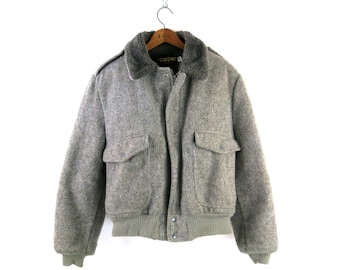 Puede incluir: Chaqueta bomber gris de lana con cuello de piel sintética, dos bolsillos delanteros y dobladillo y puños de canalé. La chaqueta está colgada de una percha de madera. La marca "Cooper" es visible en el cuello.