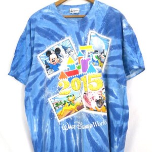 May include: Blue tie-dye t-shirt featuring Mickey Mouse, Pluto, and other Disney characters. The shirt has the text "2015" and "Walt Disney World" printed on the front. The shirt is hanging on a wooden hanger.