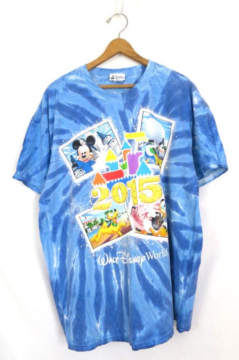 May include: Blue tie-dye t-shirt featuring Mickey Mouse, Pluto, and other Disney characters. The shirt has the text "2015" and "Walt Disney World" printed on the front. The shirt is hanging on a wooden hanger.