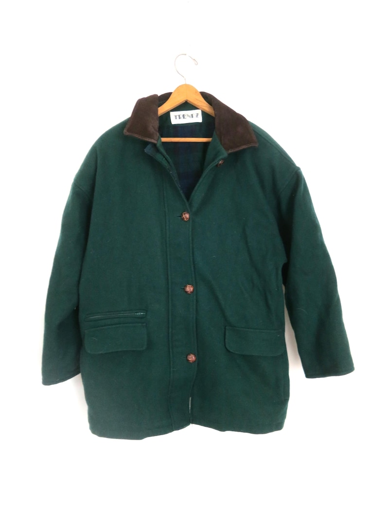 Puede incluir: Un abrigo de lana verde oscuro con cuello marr&oacute;n y botones de madera. El abrigo tiene dos bolsillos delanteros y un dise&ntilde;o cl&aacute;sico. El abrigo est&aacute; colgado de una percha de madera.