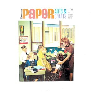 Puede incluir: Portada de libro vintage titulada "Paper Arts & Crafts" con una escena de aula. Un profesor y estudiantes se reúnen alrededor de un mural colorido. La portada tiene un esquema de color rosa y turquesa, con el precio del libro indicado en 50¢.