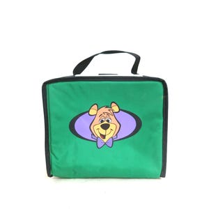 May include: Green lunch bag with a black handle and trim. The bag features a cartoon bear character in an oval shape with a purple background. The bear has a purple bow tie.