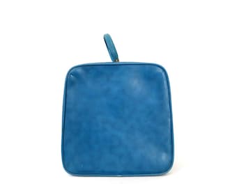 May include: A square, teal-colored handbag with a small, curved handle. The bag has a textured surface and a simple, classic design. The handle is attached to the top of the bag with a gold-colored metal piece.
