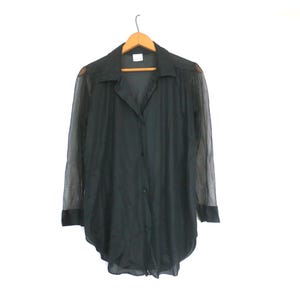Peut inclure: Une chemise noire boutonnée avec des manches transparentes est suspendue à un cintre en bois. La chemise a un col classique et un ourlet légèrement arrondi. Les manches sont en tissu transparent, et les poignets sont noirs.