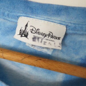May include: A close-up of a blue tie-dye t-shirt with a Disney Parks label. The label shows a castle silhouette and the words "Disney Parks Authentic Original". The shirt is on a wooden hanger.