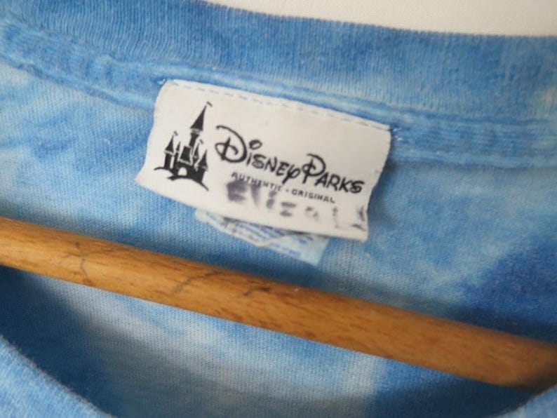 May include: A close-up of a blue tie-dye t-shirt with a Disney Parks label. The label shows a castle silhouette and the words "Disney Parks Authentic Original". The shirt is on a wooden hanger.