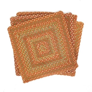 Puede incluir: Un conjunto de cuatro posavasos cuadrados trenzados. Los posavasos están hechos de un material tejido en tonos naranja, verde y crema. Los posavasos están dispuestos en un patrón ligeramente desplazado, mostrando la forma cuadrada completa.