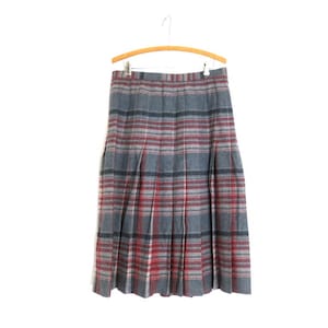 May include: A grey, red, and black plaid skirt hangs on a wooden hanger. The skirt is pleated and has a classic, vintage style. The plaid pattern is a mix of grey, red, and black lines.
