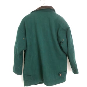Puede incluir: Un abrigo de lana verde oscuro con cuello marr&oacute;n cuelga de una percha de metal. El abrigo tiene dos bolsillos delanteros, cierres de bot&oacute;n y un dise&ntilde;o cl&aacute;sico. El abrigo es de talla mediana.