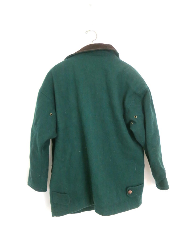 Puede incluir: Un abrigo de lana verde oscuro con cuello marr&oacute;n cuelga de una percha de metal. El abrigo tiene dos bolsillos delanteros, cierres de bot&oacute;n y un dise&ntilde;o cl&aacute;sico. El abrigo es de talla mediana.