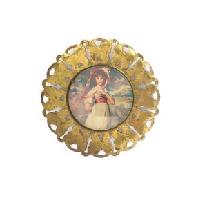 May include: A circular gold-toned metal frame featuring a decorative border of repeating butterfly silhouettes. The center contains a circular print depicting a person with dark hair wearing a light-colored dress and a pink headpiece against a cloudy sky.