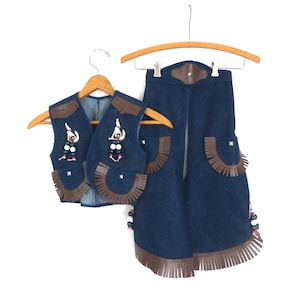 Op de afbeelding: Een denim vest en broek set met bruine lederen details. Het vest heeft decoratieve kralen en leren franjes. De broek heeft leren franjes en zakken. De set hangt aan houten kleerhangers.