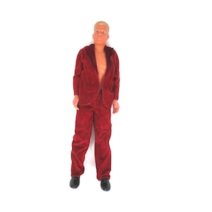 May include: A plastic action figure with short blonde hair, wearing a dark red velvet-textured blazer and matching trousers. The figure has an open jacket, revealing a bare chest, and is wearing black shoes on its feet.