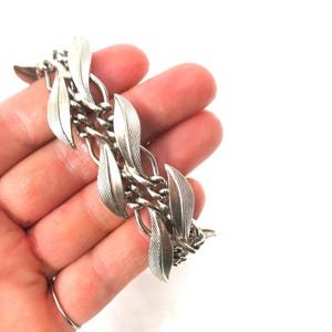 May include: Silver-toned bracelet featuring a leaf design. The bracelet is composed of interconnected leaf-shaped links and chain segments. The leaves have detailed veining, and the bracelet is held in a hand against a white background. A ring is visible on the index finger.
