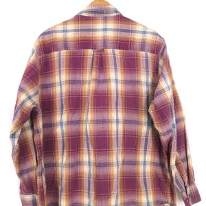 May include: A long-sleeved plaid shirt in shades of burgundy, gold, and blue, hanging on a wooden hanger. The back of the shirt is visible, showcasing the classic check pattern. The shirt has buttoned cuffs and a button-down collar.