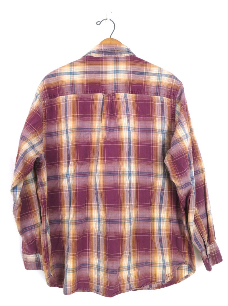 May include: A long-sleeved plaid shirt in shades of burgundy, gold, and blue, hanging on a wooden hanger. The back of the shirt is visible, showcasing the classic check pattern. The shirt has buttoned cuffs and a button-down collar.