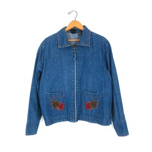 Könnte beinhalten: Eine blaue Jeansjacke mit durchgehendem Frontreißverschluss, klassischem Kragen und langen Ärmeln. Zwei aufgesetzte Vordertaschen sind mit gestickten Ahornblattmotiven in Rot und Braun verziert. Die Jacke hängt an einem hölzernen Kleiderbügel.