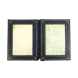 May include: An open, vintage photo album with a dark blue cover and gold decorative trim. The album has two rectangular openings for photos, one with a cream-colored background and the other with a light green patterned background.