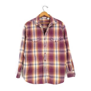 May include: A button-down plaid shirt in shades of burgundy, gold, and cream, hanging on a wooden hanger. The long-sleeved shirt features two chest pockets and a classic collar. The shirt is made of a soft, comfortable material.