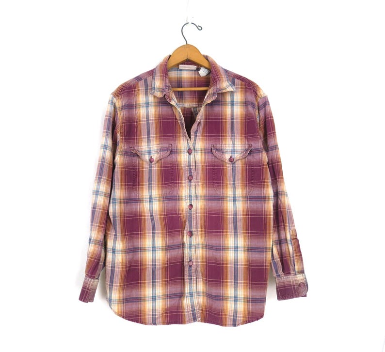 May include: A button-down plaid shirt in shades of burgundy, gold, and cream, hanging on a wooden hanger. The long-sleeved shirt features two chest pockets and a classic collar. The shirt is made of a soft, comfortable material.
