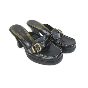 May include: A pair of black leather heeled mules with square toes. The shoes feature a gold buckle and white stitching details. The heels are approximately 3 inches high. The brand name "Mudd" is visible on the insole.