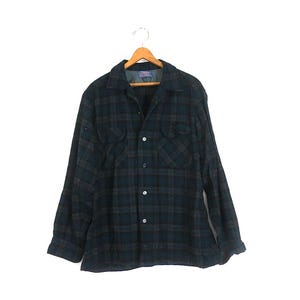 Puede incluir: Una camisa de manga larga con botones y un patrón de cuadros en color verde azulado oscuro y negro. La camisa incluye un cuello estándar, dos bolsillos en el pecho con solapas abotonadas y una fila vertical de botones. Está en una percha.