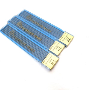 Puede incluir: Tres paquetes azules de minas de lápiz Staedtler Mars. Cada paquete tiene el nombre de la marca en letras doradas. Los paquetes están etiquetados como "2H" y "Mars Lumograph". Las minas de lápiz son visibles a través del plástico transparente.