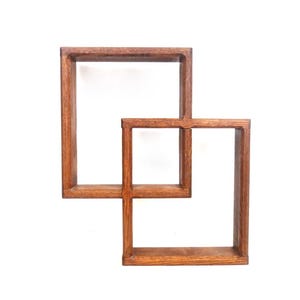 May include: A wall-mounted shelving unit constructed from two interlocking rectangular wooden frames. The frames feature a medium-brown wood grain finish and are joined together to create a geometric, open-box display structure.