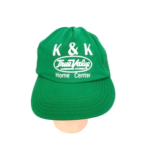 May include: A green baseball cap with a white mesh back. The cap features the text "K & K True Value Hardware Stores Home Center" in white lettering. The cap is a solid green color with a curved brim.