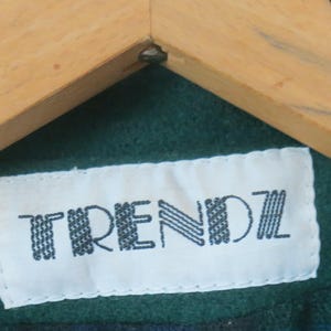 Puede incluir: Primer plano de una etiqueta blanca con la palabra "TRENDZ" en negro, cosida sobre una tela verde. La etiqueta est&aacute; debajo de una percha de madera.