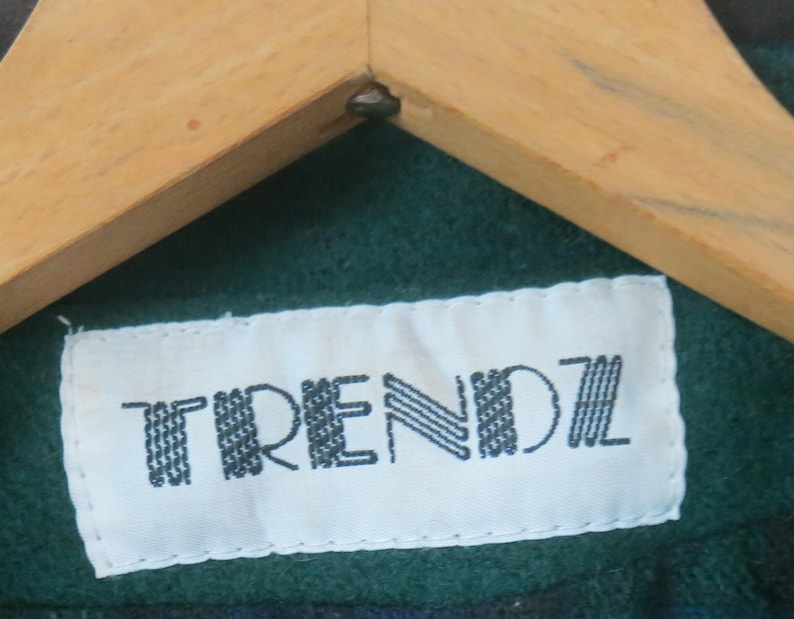 Puede incluir: Primer plano de una etiqueta blanca con la palabra "TRENDZ" en negro, cosida sobre una tela verde. La etiqueta est&aacute; debajo de una percha de madera.