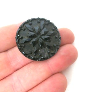 May include: A round, black button featuring a central star-like pattern with faceted, diamond-shaped segments. The outer edge of the button is decorated with a raised, scalloped border and small, evenly spaced circular dots around the circumference.