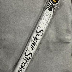 May include: A white golf putter grip with a black spider and web design. The grip has the words "Super Stroke" printed in black. The grip is in a clear plastic wrapper.