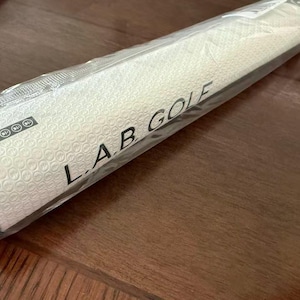 May include: A white golf club grip with a textured surface and the text "L.A.B. GOLF" printed in black. The grip is packaged in clear plastic. The grip is designed for golf clubs.