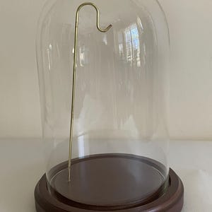 May include: A clear glass cloche with a dark brown wooden base and a gold-coloured metal hook. The glass dome sits on a circular wooden base, creating a display case. The hook is centered inside the dome.