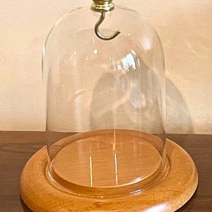 Puede incluir: Una campana de cristal transparente con un asa de latón y una base de madera. La base es de color marrón claro y de forma circular. La campana está diseñada para proteger y exhibir objetos.