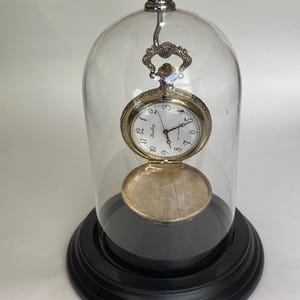 May include: A vintage pocket watch with a silver-colored case and white face, displayed inside a clear glass dome. The watch is open, revealing the inner workings. The dome sits on a black, tiered base, creating a classic decorative piece.