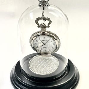 May include: A silver pocket watch, marked "GENEVA", is showcased under a clear glass dome. The watch is open, revealing a patterned interior. The dome rests on a black, tiered base, creating a decorative display. The watch is a classic timepiece.
