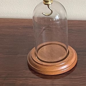 Könnte beinhalten: Eine transparente Glasglocke mit Messinghaken und -knauf, auf einem runden Holzsockel. Der Sockel hat eine warme braune Farbe, und die Glaskuppel ist leer. Die Glocke ist für die Präsentation kleiner Gegenstände konzipiert.