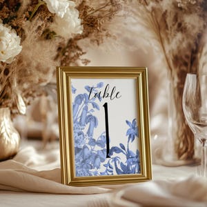 May include: A framed table number one, with a gold frame. The artwork features blue floral designs and the word "Table" in script. The number one is in a bold black font. The background includes floral arrangements and glassware.