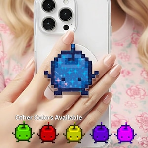 May include: A blue pixelated phone grip featuring a cute character design with star accents. The grip is attached to a white phone. The image also shows a variety of other color options available.