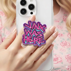 May include: A purple and blue phone grip with the text "Jinx Was Here" in a graffiti-style design. The phone accessory is attached to a clear phone case.