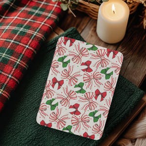 Puede incluir: Un posavasos rectangular blanco roto con un patrón repetido de lazos rojos y verdes. Los lazos tienen cintas a rayas rojas y blancas. El posavasos está sobre una superficie de punto verde oscuro, con una manta a cuadros roja y verde.