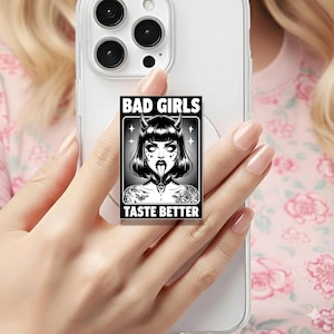 May include: A black and white phone grip with a graphic of a woman with devil horns and the text "BAD GIRLS TASTE BETTER". The image is set against a clear phone case.