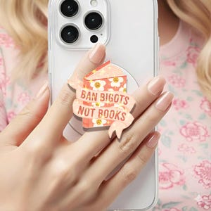 May include: A phone grip featuring a book design with the text "BAN BIGOTS NOT BOOKS" on a banner. The book has a floral pattern and sad-faced flower graphics. The grip is attached to a clear phone case.