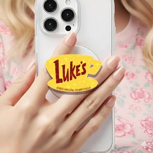 Puede incluir: Un soporte de teléfono amarillo con el texto "Luke's" en rojo, con forma de taza de café. El soporte también incluye el texto "Stars Hollow, Connecticut". El soporte está adherido a una funda transparente.