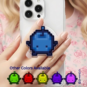 May include: A blue pixelated phone grip is attached to a clear phone case. The grip has a cartoon character design. Below the grip, the text "Other Colours Available" is displayed, with examples in green, red, yellow, purple, and pink. The word "CUSTOM" is written vertically.